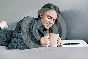 Autumn winter portrait of young girl in warm knitted sweater at home on sofa with notebook and cup of hot drink