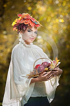 Autumn Woman. Beautiful creative makeup and hair style in outdoor shoot . Girl with leaves in hair holding a basket with apples