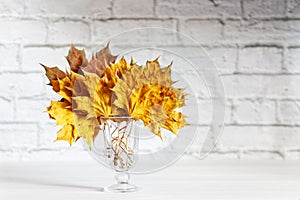 Autumn yellow leaves in transparent glass on white wooden table. Shallow focus.