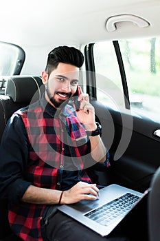 Always available. Handsome young indian man working on his laptop and talking on the phone while sitting in the car