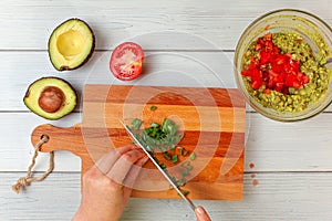 Avocado halves, tomato, hand slicing spring onion - basic guacamole ingredients on working board, view from above