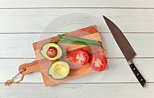 Avocado halves, tomato, spring onions - basic guacamole ingredients on working board, knife near, view from above