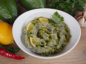 Avocado puree in a white bowl, lemon, garlic, chili pepper, herbs on a light wooden table closeup. Traditional delicious recipe