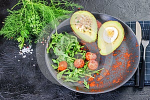 Avocado stuffed with eggs, tuna and sauce, arugula and dill on the black modern dish. Black cutlery, napkin and background. Keto