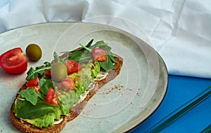 Avocado toast of rye bread with sauce, tomatoes, arugula on large grey plate on blue background. Vegetarian useful breakfast.