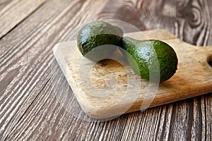Avocados on natural pine cutting board on wooden background. Two whole green fresh tropical fruits on brown table