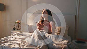 Awake woman typing phone in morning sunlight. Smiling calm girl resting in bed