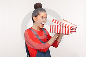 Awesome holiday gift! Portrait of surprised nosy pretty girl with hair bun in denim overalls looking into present box