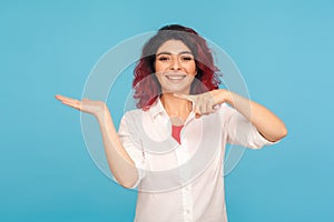 Awesome joyful hipster woman with fancy red hair presenting advertising area on her palm and pointing to copy space