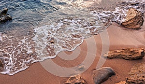 Awesome nature background. Soft beautiful ocean wave on sandy beach with rocks during sunset. Backdrop