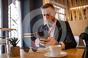 Awesome young guy or businessman reading news on smartphone and drinking morning coffee from big white cup