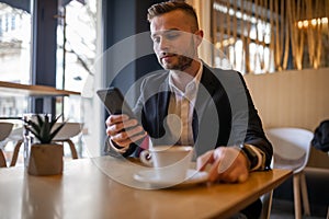 Awesome young guy or businessman reading news on smartphone and drinking morning coffee from big white cup