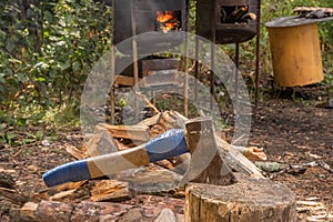 Ax stuck into a wooden block against the background of firewood and a stove with fire for outdoor cauldrons