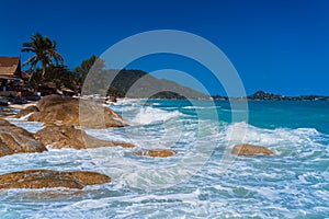 Azure Ocean waves roll round stones on the sandy beach of Lamai, Koh Samui, Thailand. Palm trees and bungalows.