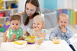 Babies having lunch in kindergarten. Kids eat healthy food in creche. Carer with children sitting at dinner table in