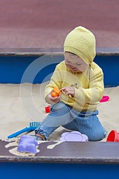 Baby 10 months playing in the sandbox. Child girl boy in a yellow knitted sweater plays with sandbins, vertical portrait Caucasian