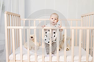 A baby of 8 months stands in a crib with toys in pajamas in a bright children`s room after sleeping and looks at the camera, a