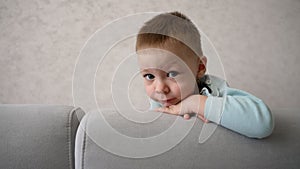 The baby appears from the sofa. A child plays hide and seek. A little boy is hiding behind the sofa. A little boy looks