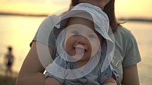 A baby in the arms of her mother smiles at the river at sunset. A six-month-old child in a panama hat smiles cheerfully