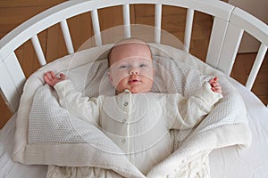 The baby in a beige bodysuit and beige envelope lies on his back on white bed linen in a white round crib