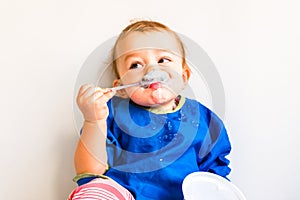 Baby with bib enjoying while eating a yogurt with spoon and stains