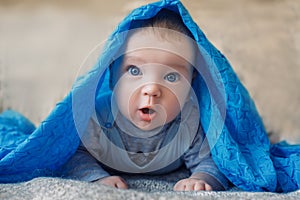 Baby with big blue eyes looks in surprise at the camera from under the blue fabric