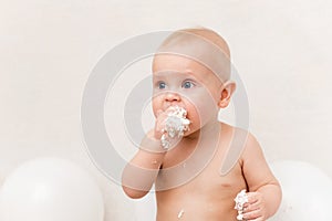 Baby birthday party. Infant eating birthday cake. The boy on a light background with white ballons and copy space smash the cake