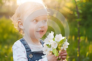 Baby with bouquet of flowers in the garden in sunlight. Cute happy summer blond girl in the garden. Apple blossom