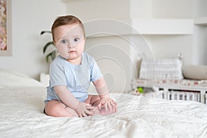 Baby boy 6 months old in blu bodysuit smiling and sitting on white bed at home