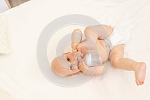 Baby boy 8 months old lying in diapers on a white bed with a bottle of milk at home legs up, top view, baby food concept, baby