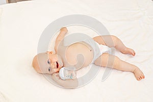 Baby boy 8 months old lying in diapers on a white bed with a bottle of milk at home legs up, top view, baby food concept, baby