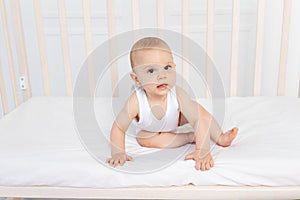 Baby boy 8 months old sitting in a crib in a children`s room in white clothes and looking away, baby`s morning, baby products