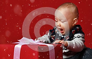 Baby boy with big blue eyes wearing warm sweater sitting in front of his present in wrapped box with ribbon over red background.