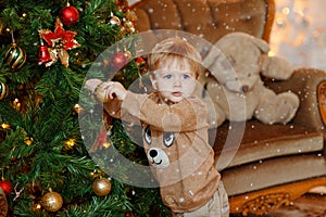 Baby boy blonde in beige sweater dresses up Christmas tree, hang