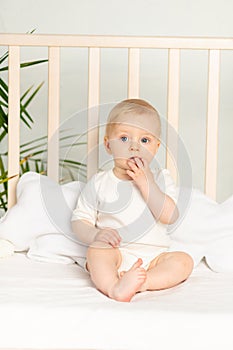 Baby boy blonde with blue eyes in a white bodysuit on a bed with cotton underwear at home before going to bed sucks a finger
