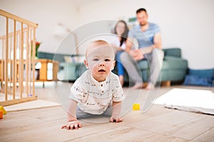 A baby boy crawling on the floor at home, parents in the background.