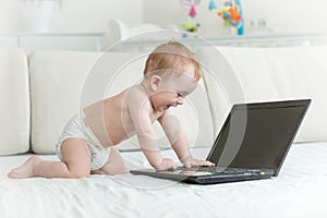 Cute baby boy in diapers crawling on bed towards laptop