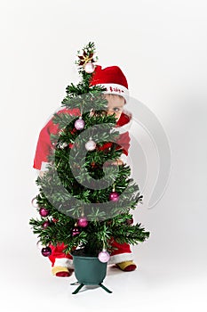 Baby boy dressed as Santa Claus hiding behind Christmas tree