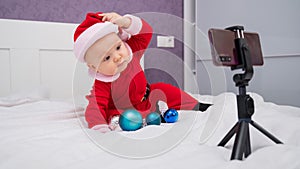 A baby boy dressed in a Santa suit sits on the bed and looks at the sick phone. Christmas greetings by video call