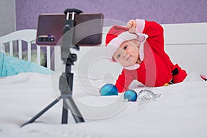 A baby boy dressed in a Santa suit sits on the bed and looks at the sick phone. Christmas greetings by video call