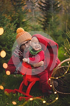 Baby boy with father in the red knit jumpsuit and Santa hat smiling and holding rocking-horse with pine trees on the background