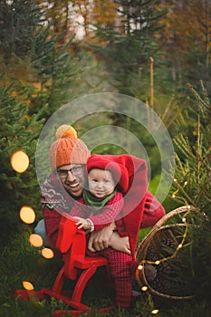 Baby boy with father in the red knit jumpsuit and Santa hat smiling and holding rocking-horse with pine trees on the background