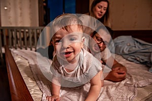 Baby boy lies on stomach on bed, smiling at camera. Family efocused from behind. Care of children