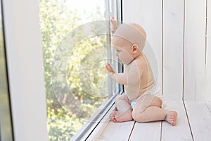 Baby boy 8 months old sitting in diapers on the window sill and looking into the distance, a place for text