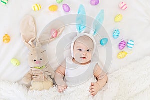 A baby boy with rabbit ears on his head is lying in a crib with a bunny toy and Easter eggs, a cute funny smiling little baby.