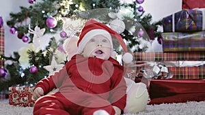 Baby boy in a red Santa costume is lying under the Christmas tree.