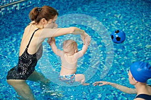 Baby boy running to ball inflatable toy. Coach raising child upright to perform exercise with hands up and feet in water