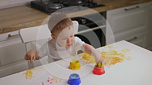 Baby boy sitting behind table and drawing with fingers covered in colorufl paint