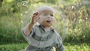 Baby boy sitting on green grass outdoor playing with soap bubbles slow motion Full HD