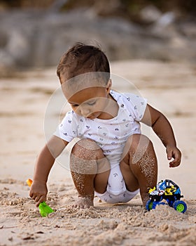 Baby boy sitting on sandy beach, playing with sand scoop and toy car. Warm sunny day. Happy childhood. Summer vacation at the sea